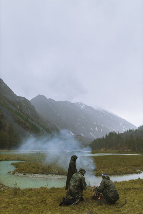 Free Two people camping by a river in a mountainous area with smoke rising from a campfire. Stock Photo