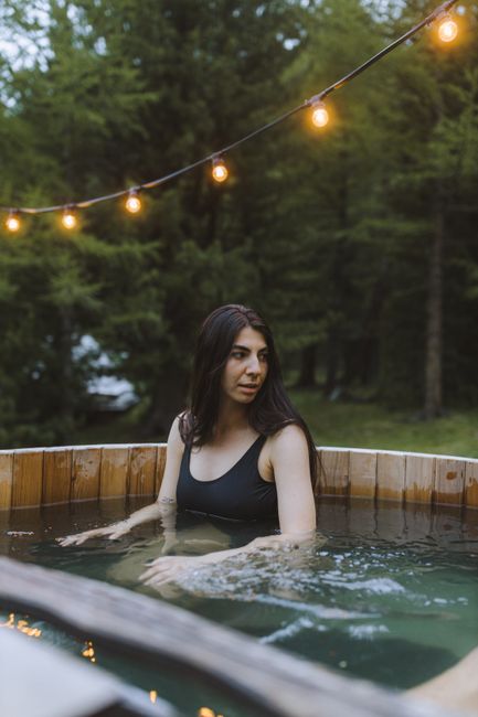 A woman enjoying a peaceful outdoor soak in a hot tub, surrounded by glowing string lights and lush greenery.