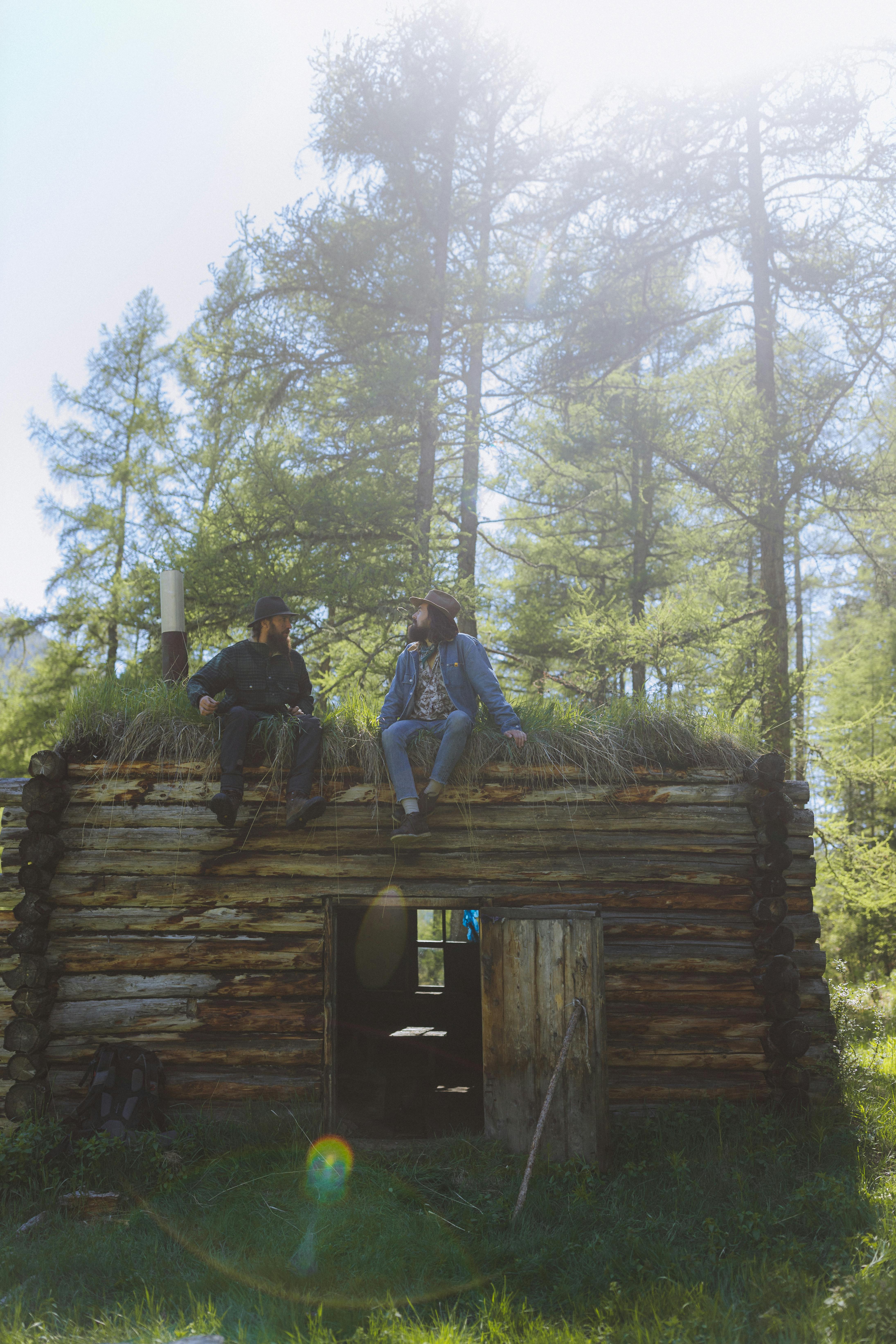Two adults enjoy a conversation sitting on a cabin roof surrounded by lush greenery.