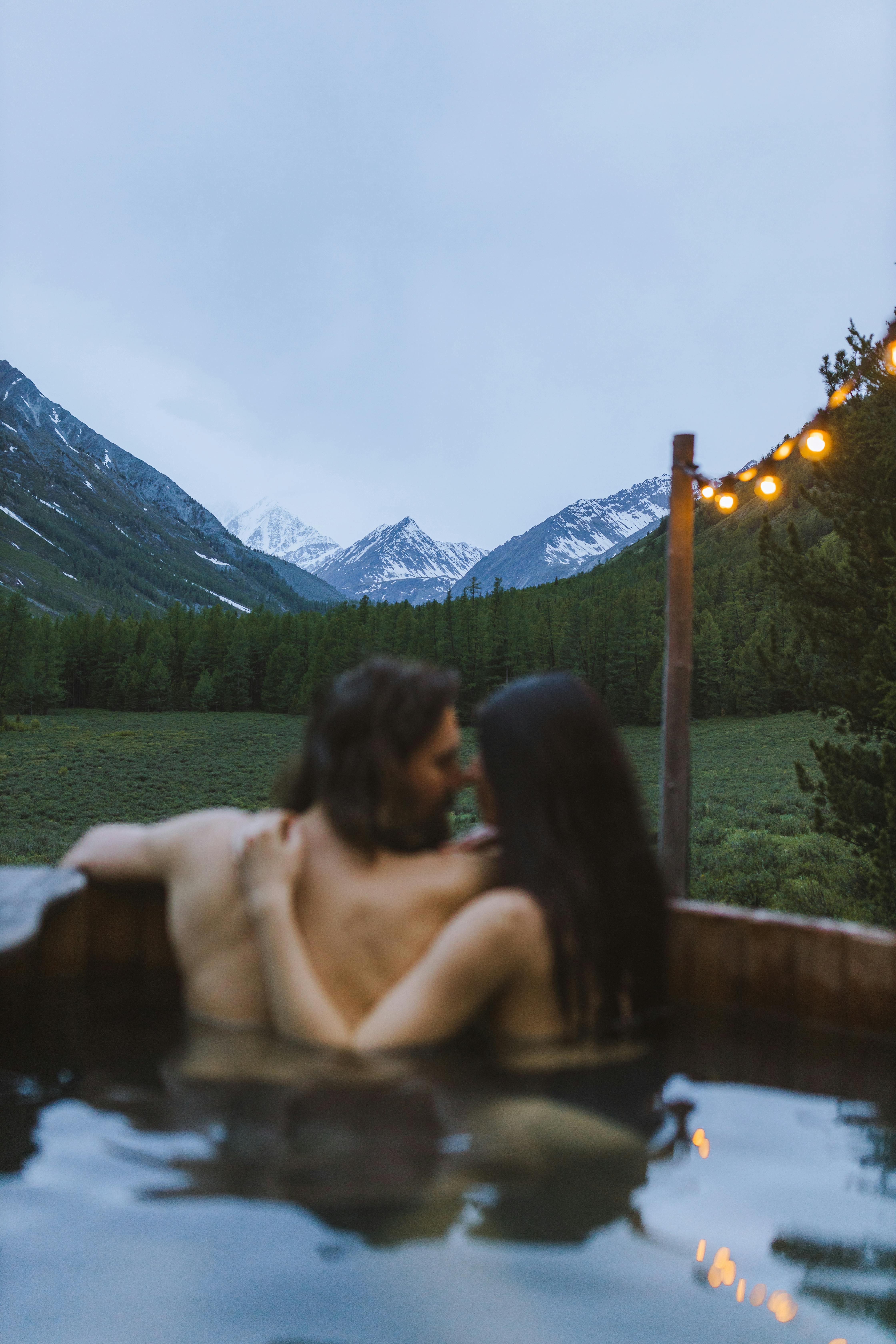 Romantic couple embracing in an outdoor hot tub, surrounded by majestic mountains at twilight.