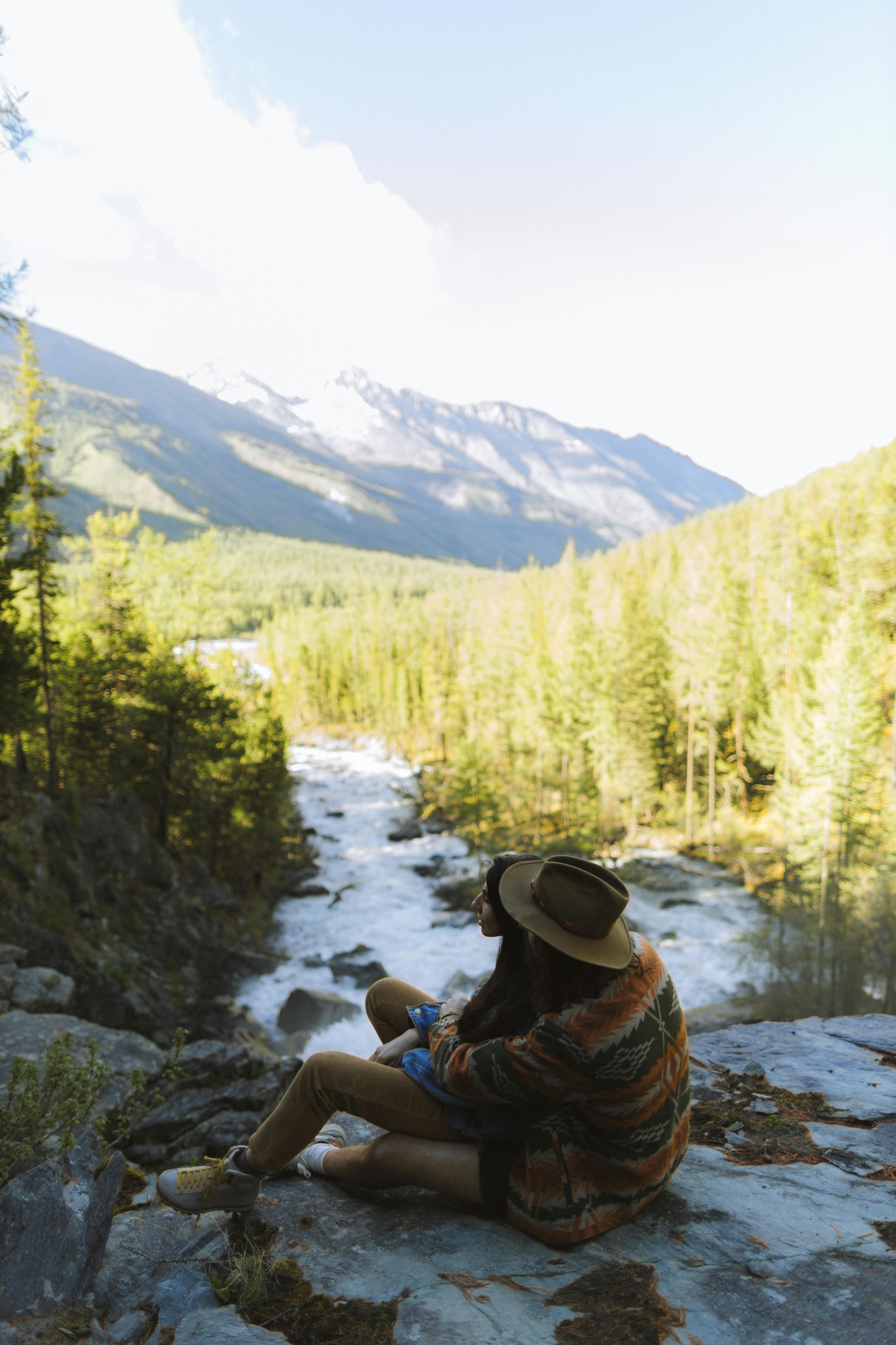 Romantic couple sits on a cliff overlooking a river and mountains, enjoying the serene landscape.