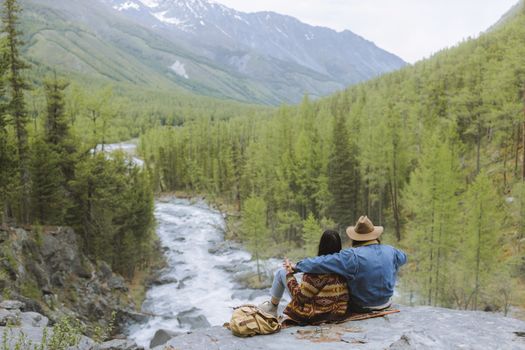 A couple sits on a rock, enjoying the natural scenery of mountains and forest by a river.