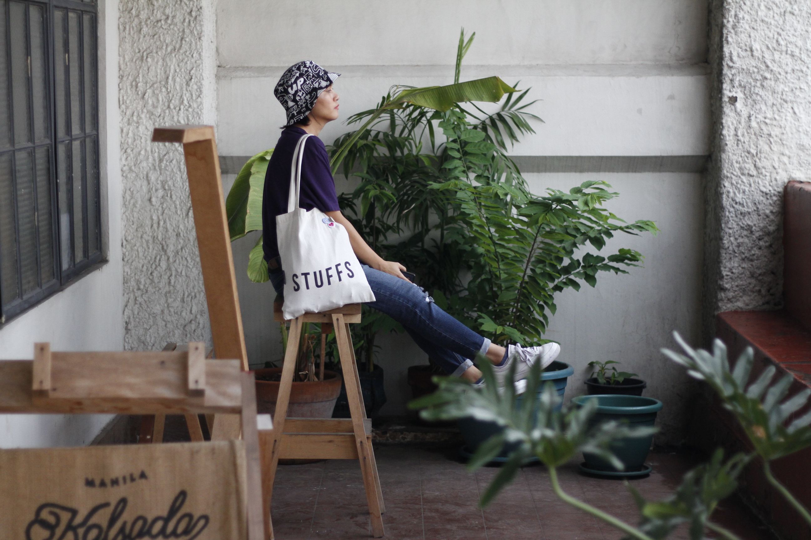 A woman sitting indoors on a wooden chair with a casual tote bag surrounded by plants.