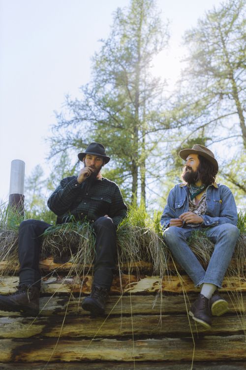 Free Two men sitting on a log, enjoying a sunny day in a park surrounded by trees. Stock Photo
