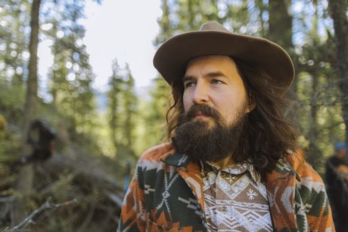 Free A bearded man in a patterned jacket and hat stands in a sunlit forest. Stock Photo
