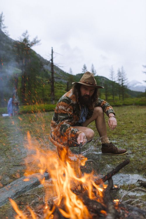 Free Man with a hat sitting by a campfire in scenic outdoor wilderness, surrounded by nature. Stock Photo