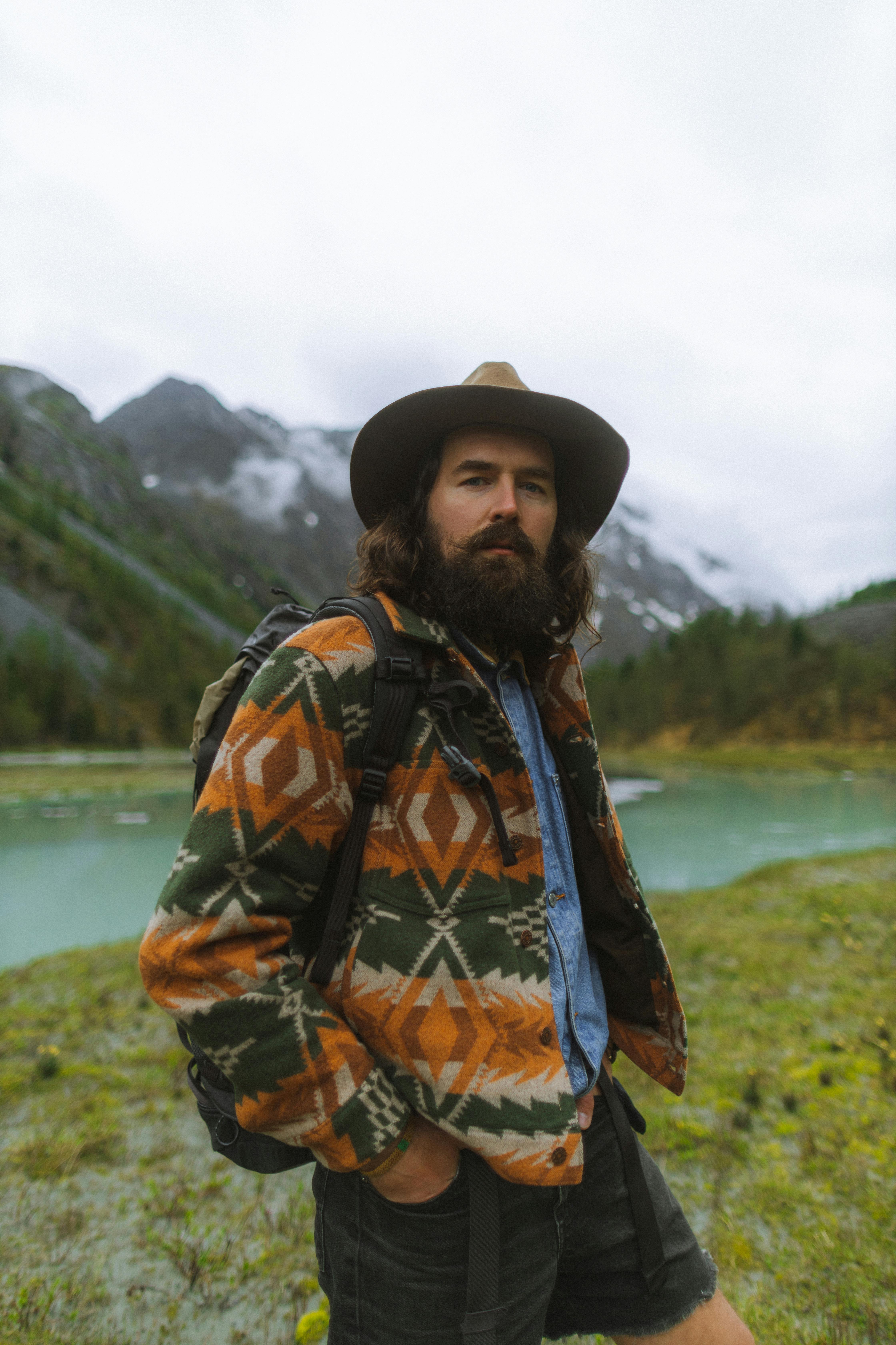 bearded man in hat and patterned jacket standing near lake with hands in pockets