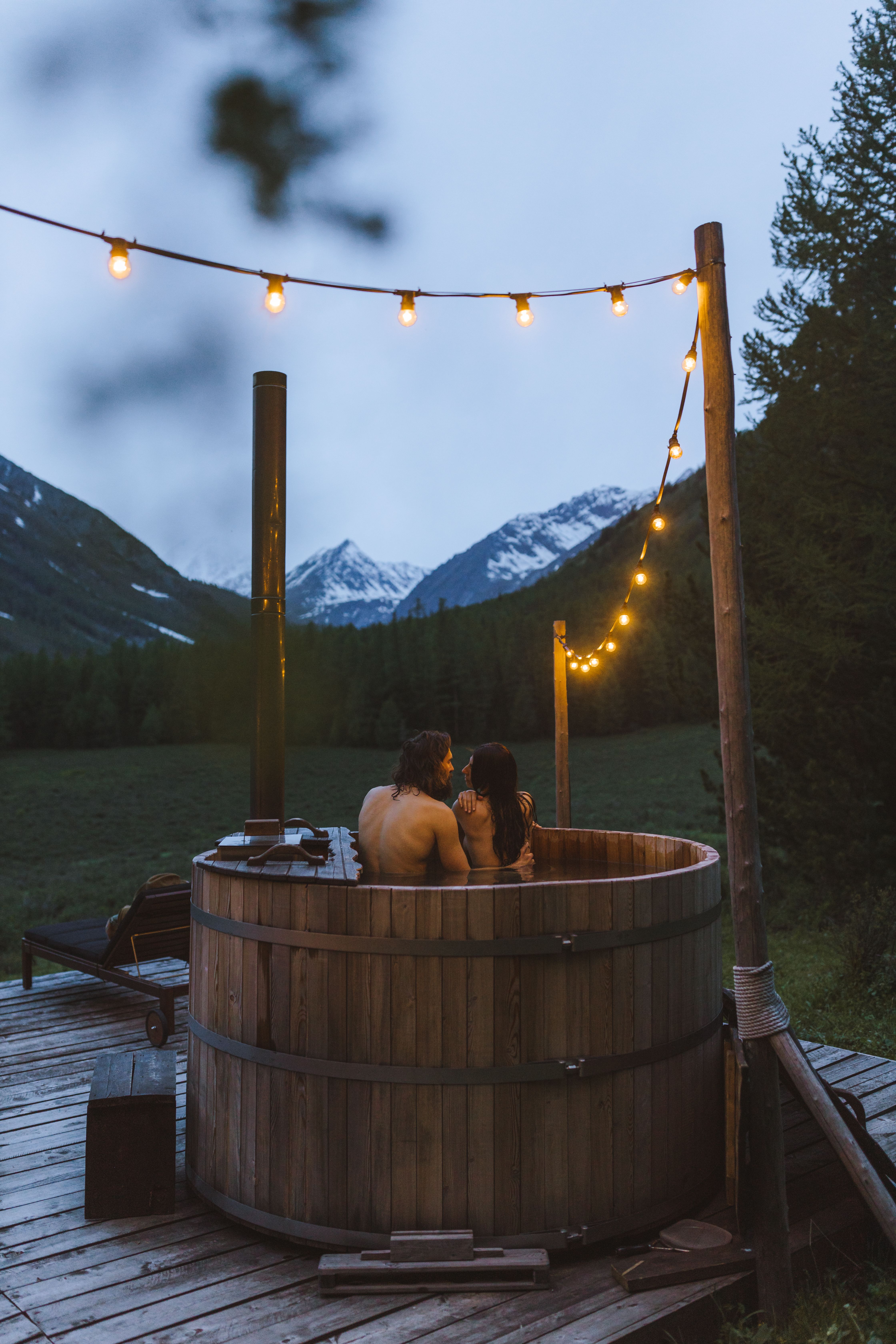 A couple in a wooden hot tub under string lights at dusk