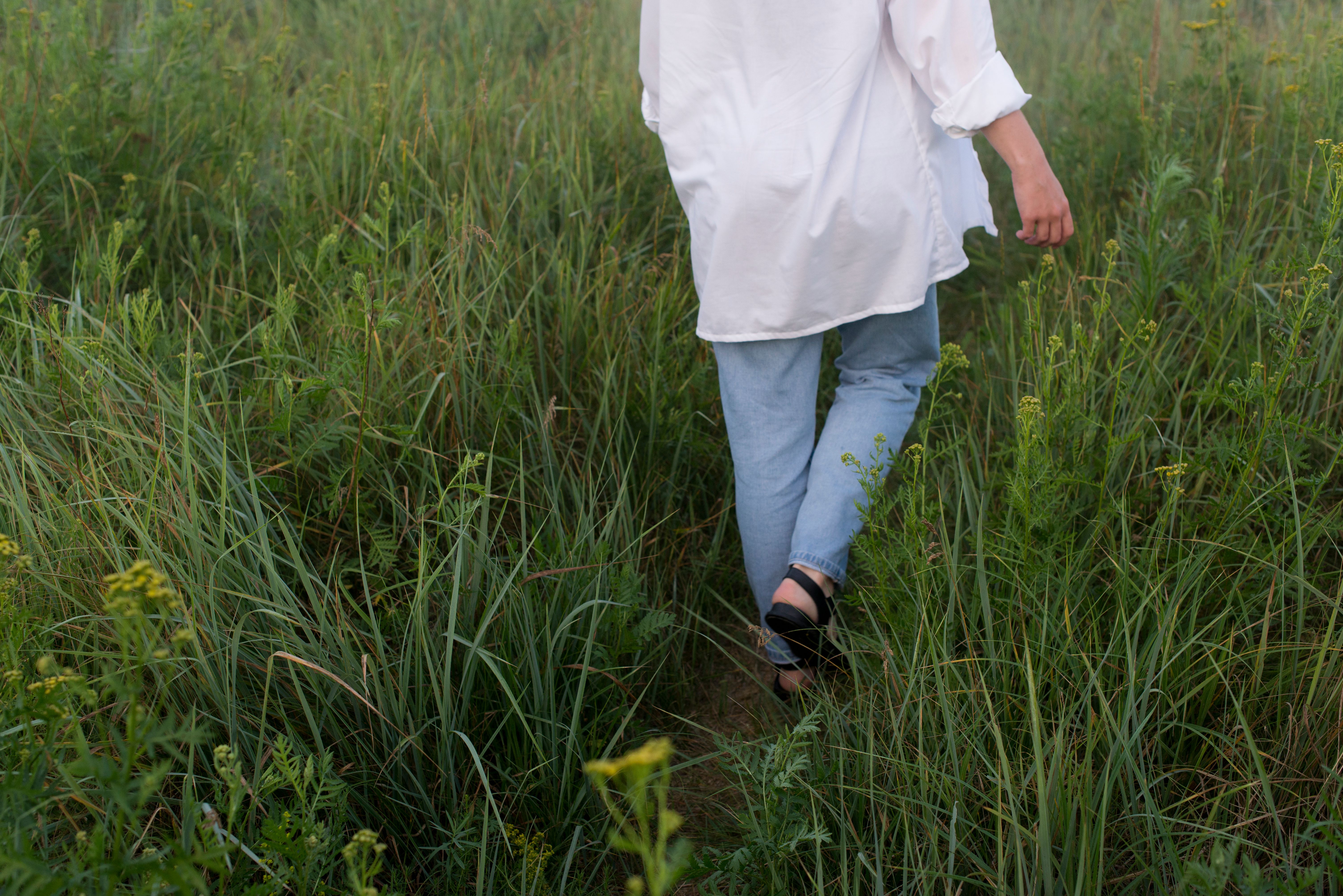 A woman in denim pants and white long sleeves walking through a lush grassy field, captured from behind.