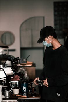 A barista in a café using a coffee machine, wearing a cap and mask.