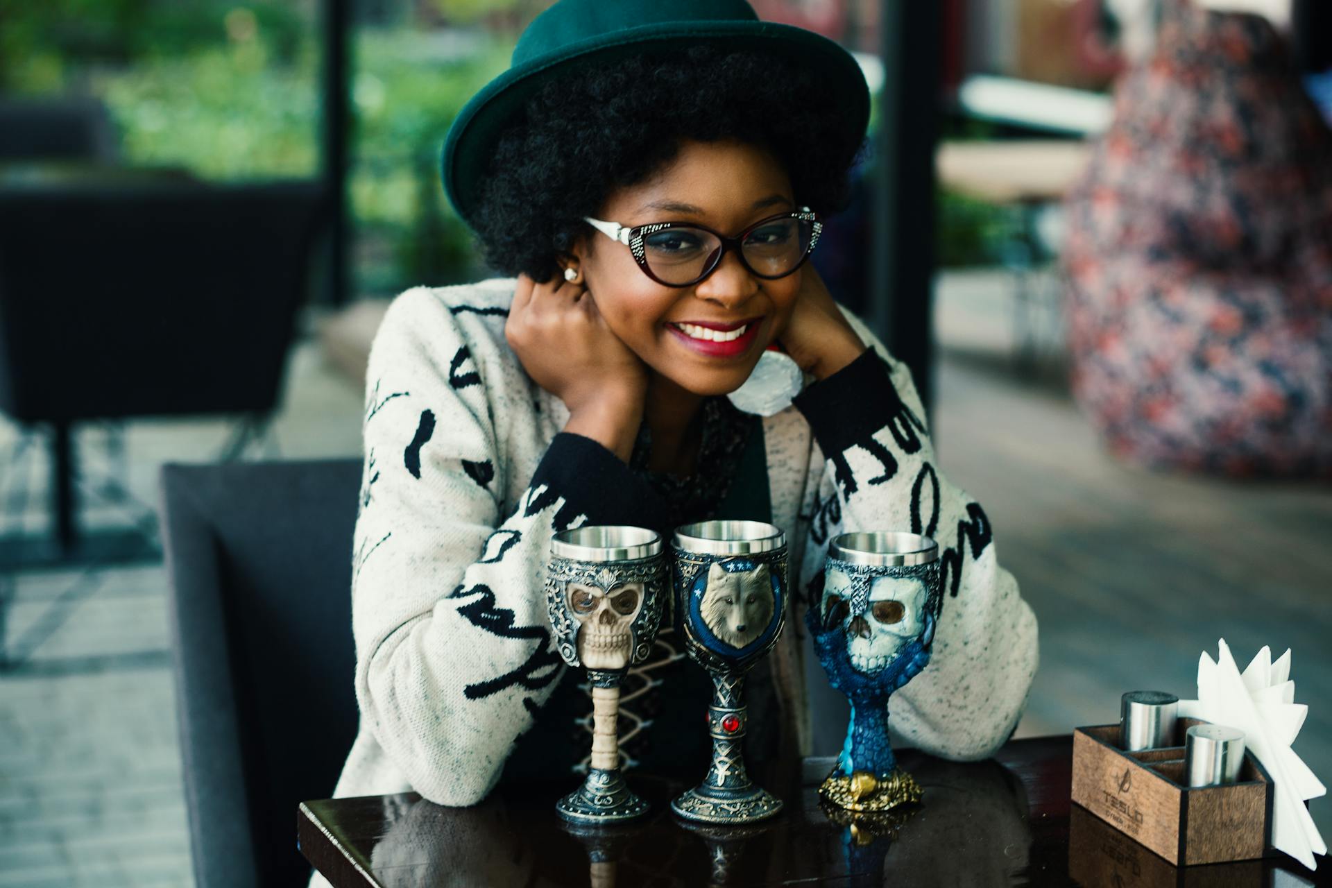 Smiling woman in glasses at an outdoor café with ornate goblets. Stylish and engaging lifestyle image.