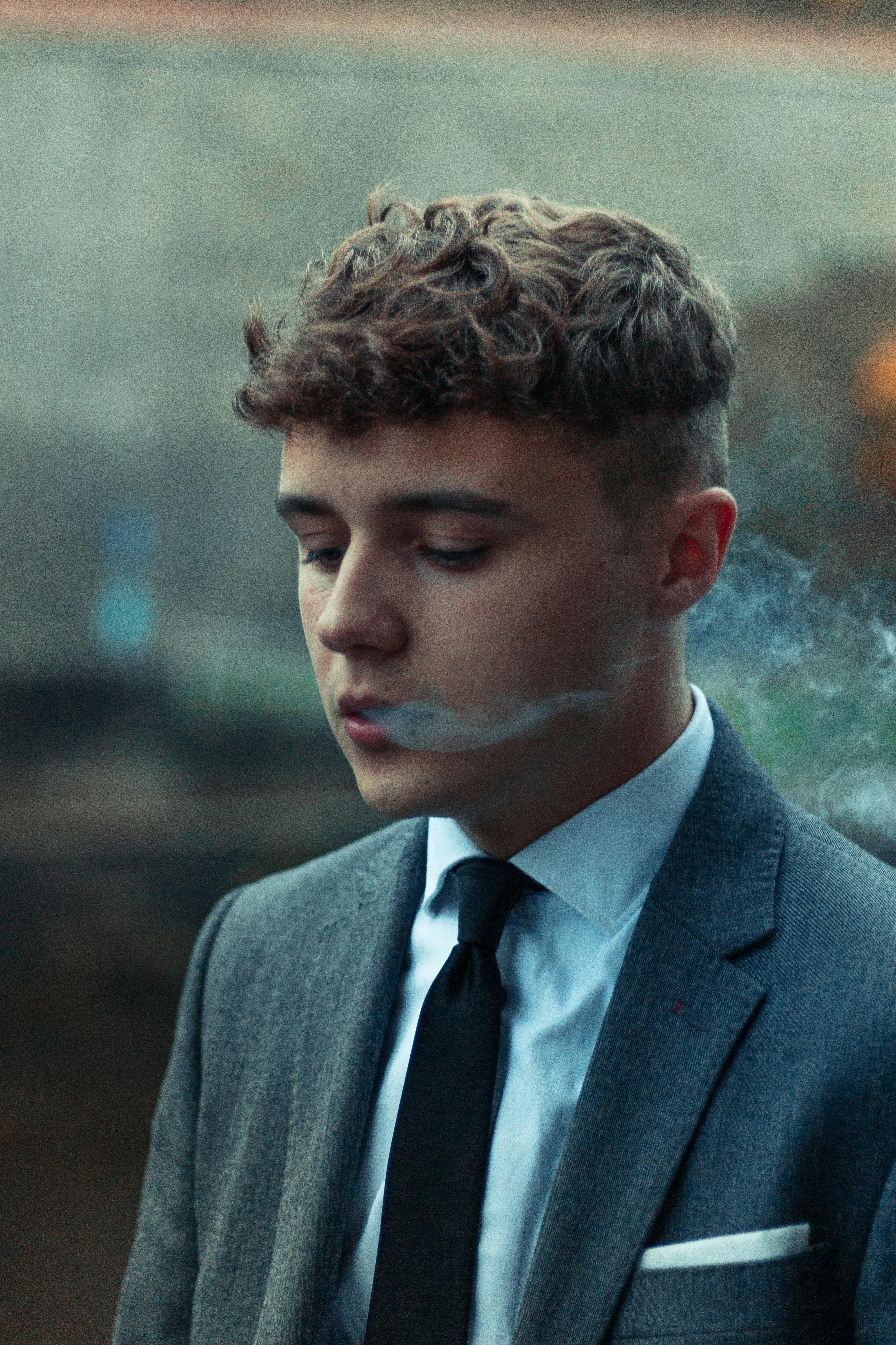 A young man with curly hair wearing a suit and tie, exhaling smoke.