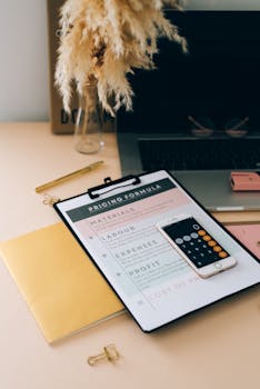 Organized desk setup with a clipboard, laptop, and smartphone displaying a pricing formula.
