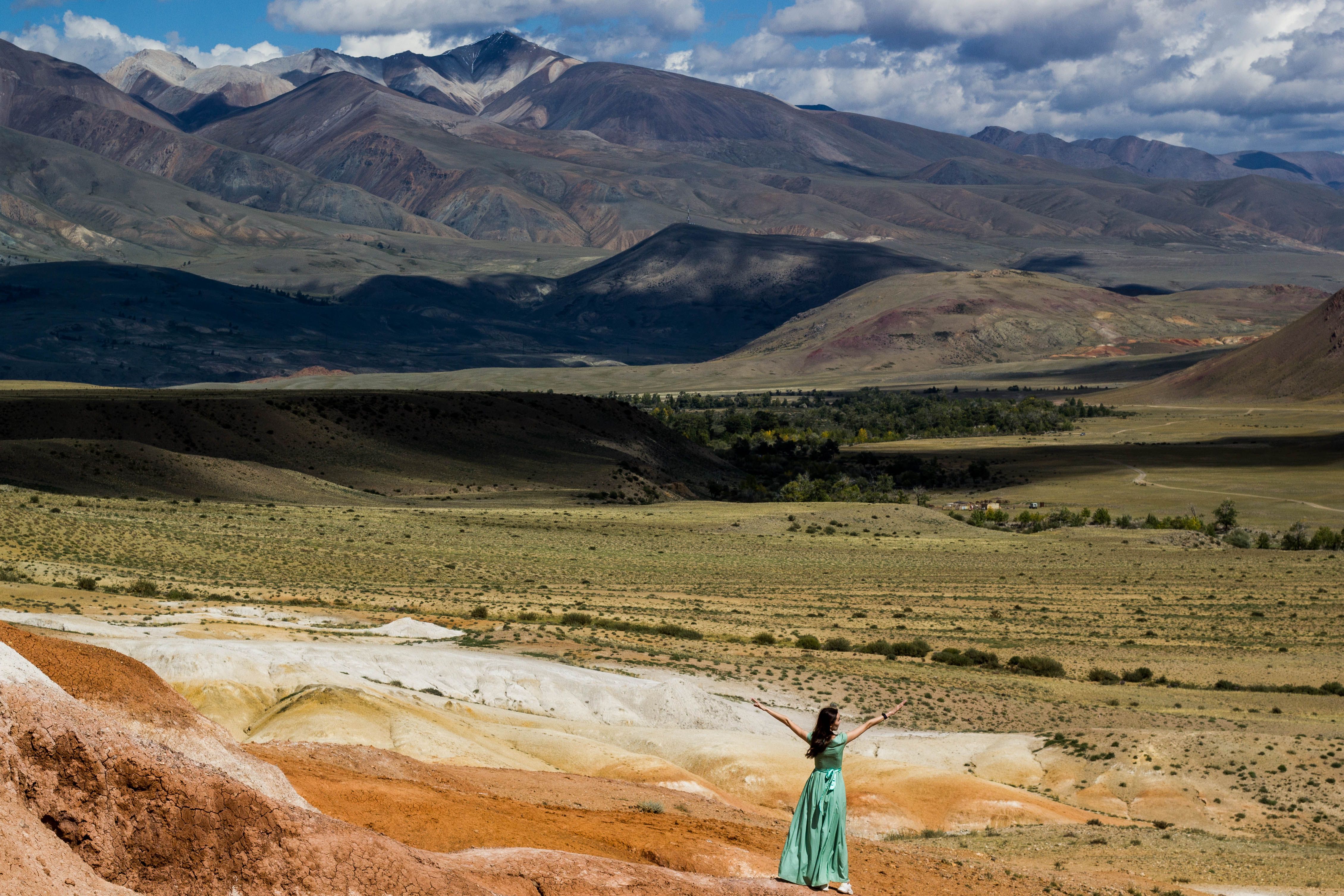 Woman in green dress stands with open arms, admiring the scenic Altai Mountains landscape.