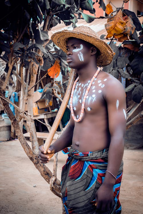 Side View Shot of a Man with White Paint on His Chest Wearing a Straw Hat while Holding a Thick Stick