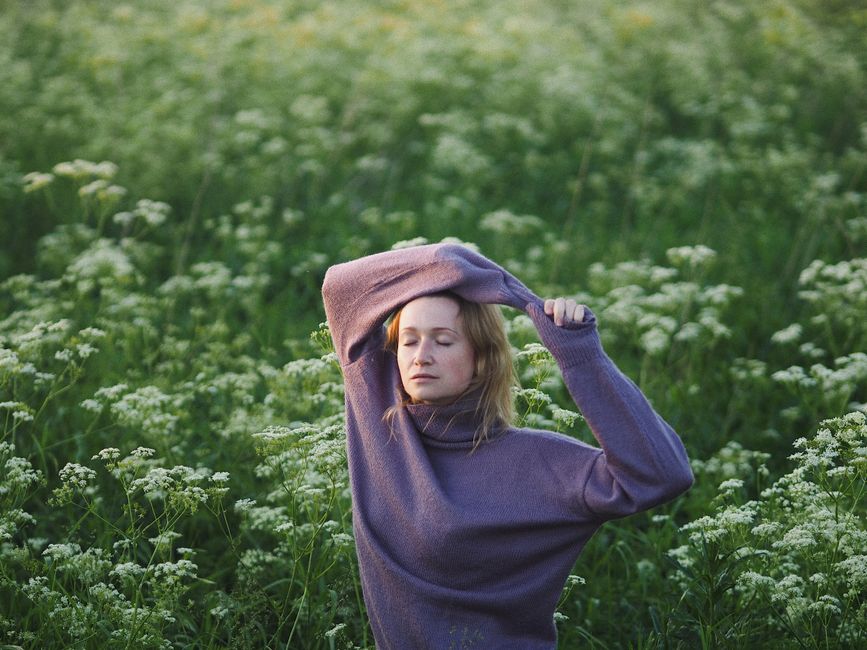 A serene woman with eyes closed in a purple sweater poses in a vibrant green meadow.