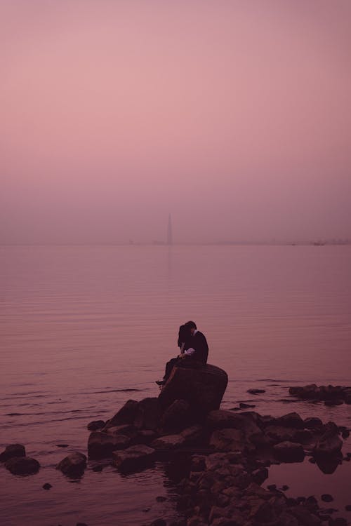 Couple Sitting on the Rock Beside Water During Sunrise