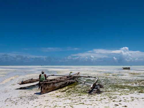 Free A person sitting on a rustic boat during low tide on a Zanzibar beach with a clear blue sky. Stock Photo