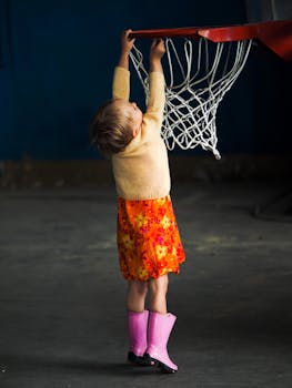 A young child in a floral skirt and pink boots reaching up to touch a basketball hoop indoors.