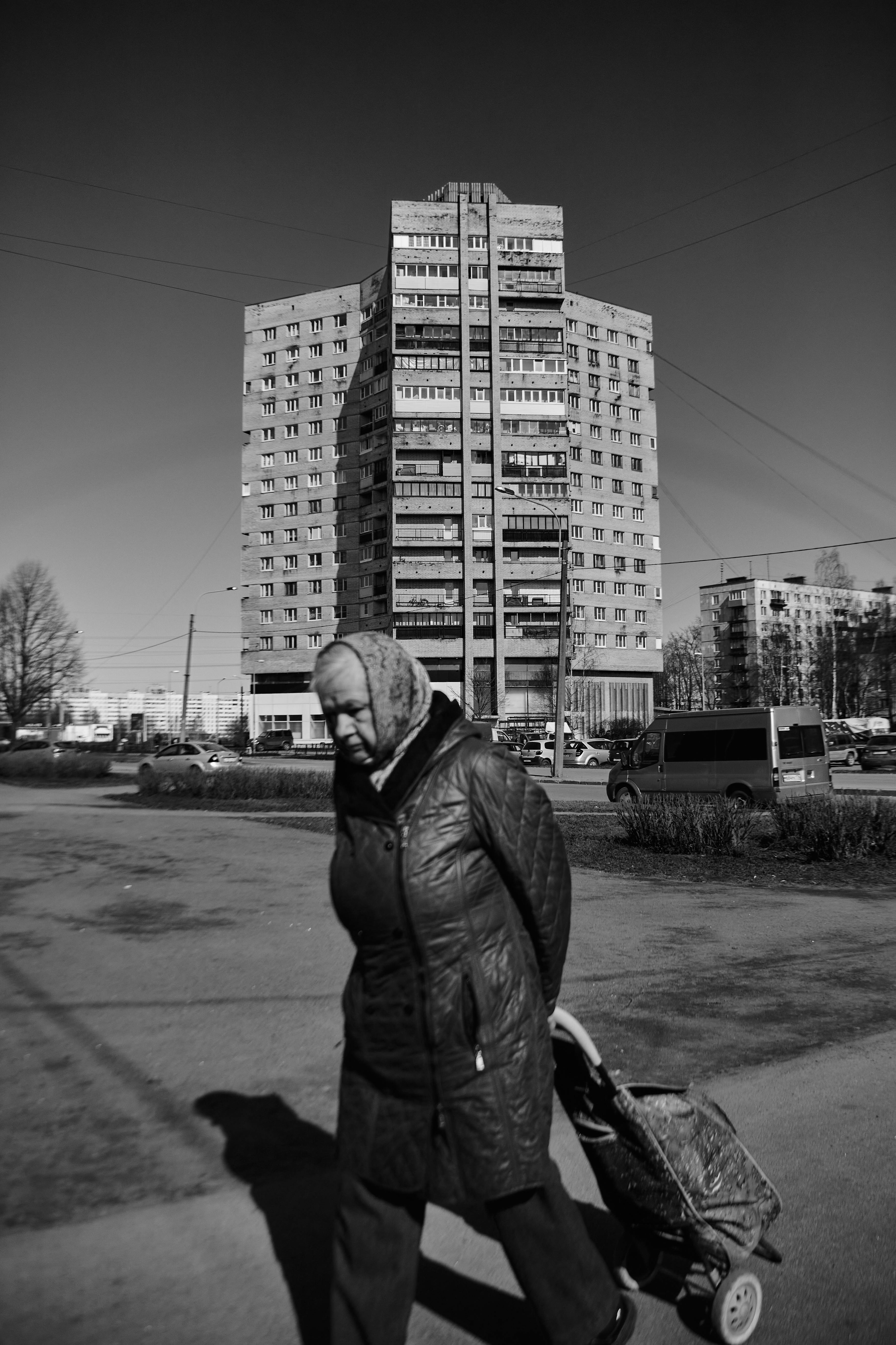 Black and white photo of an elderly woman walking with a trolley in a cityscape in Saint Petersburg, Russia.