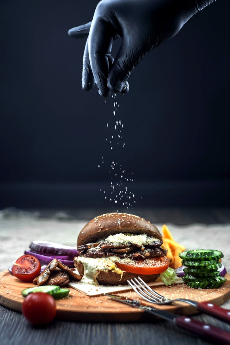 Person's Hand In Black Latex Glove Salting Burger On Wooden Chopping Board