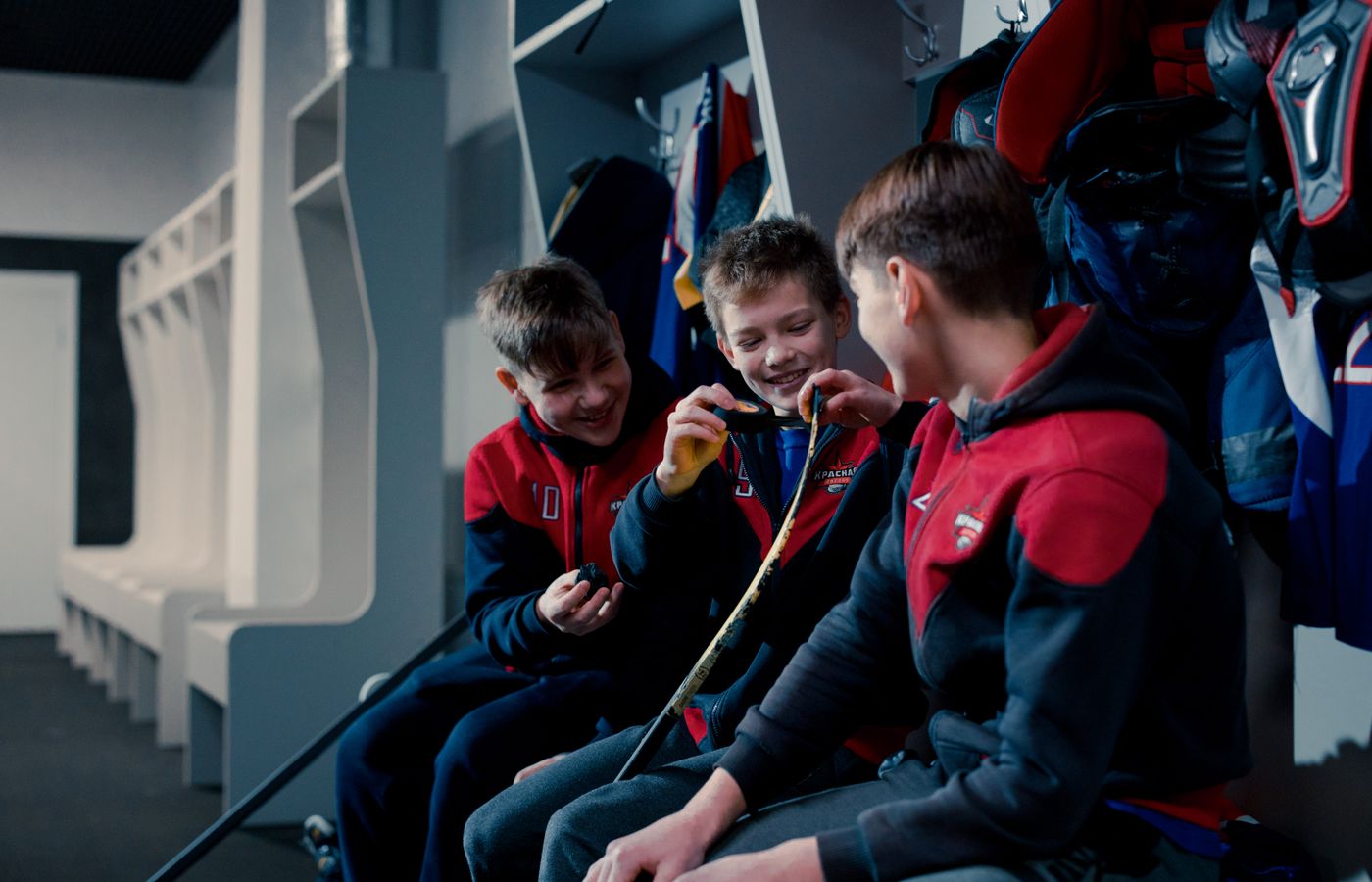 Three young boys in hockey gear taping sticks and laughing in a locker room.