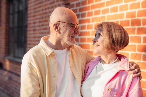 Free Elderly couple smiling and hugging against a sunlit brick wall, enjoying a moment of love. Stock Photo