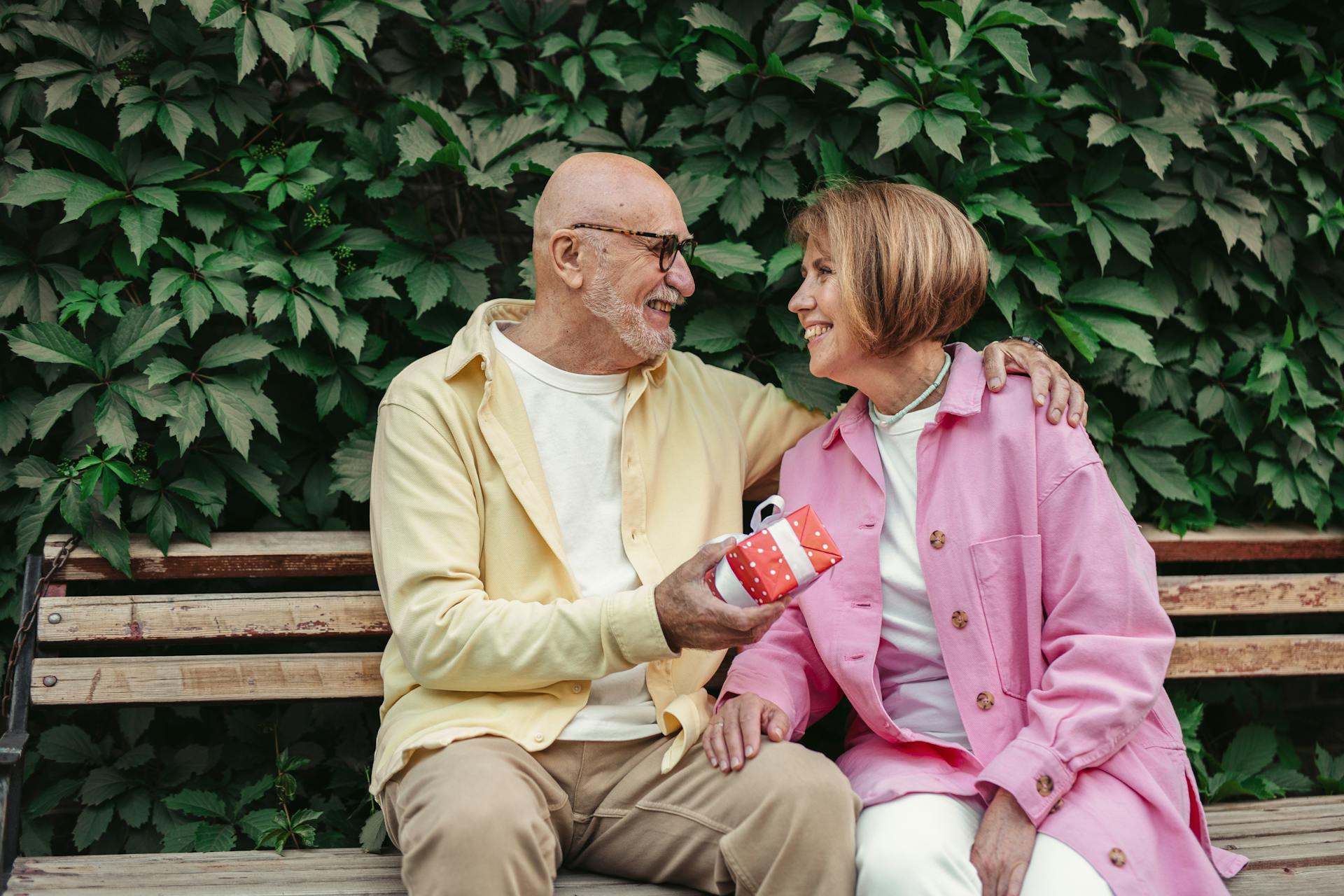 A senior couple sits on a bench exchanging a gift, expressing affection and happiness.