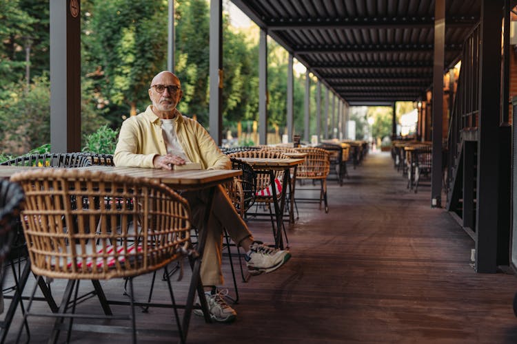 Elderly Man In Yellow Shirt And Eyeglasses Sitting On Wooden Chair On Patio