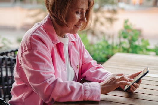Elderly woman in pink jacket using a smartphone at outdoor wooden table.