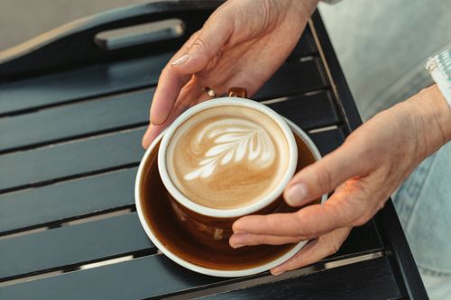 Free A close-up of a latte with intricate art on a wooden table, held by hands. Stock Photo