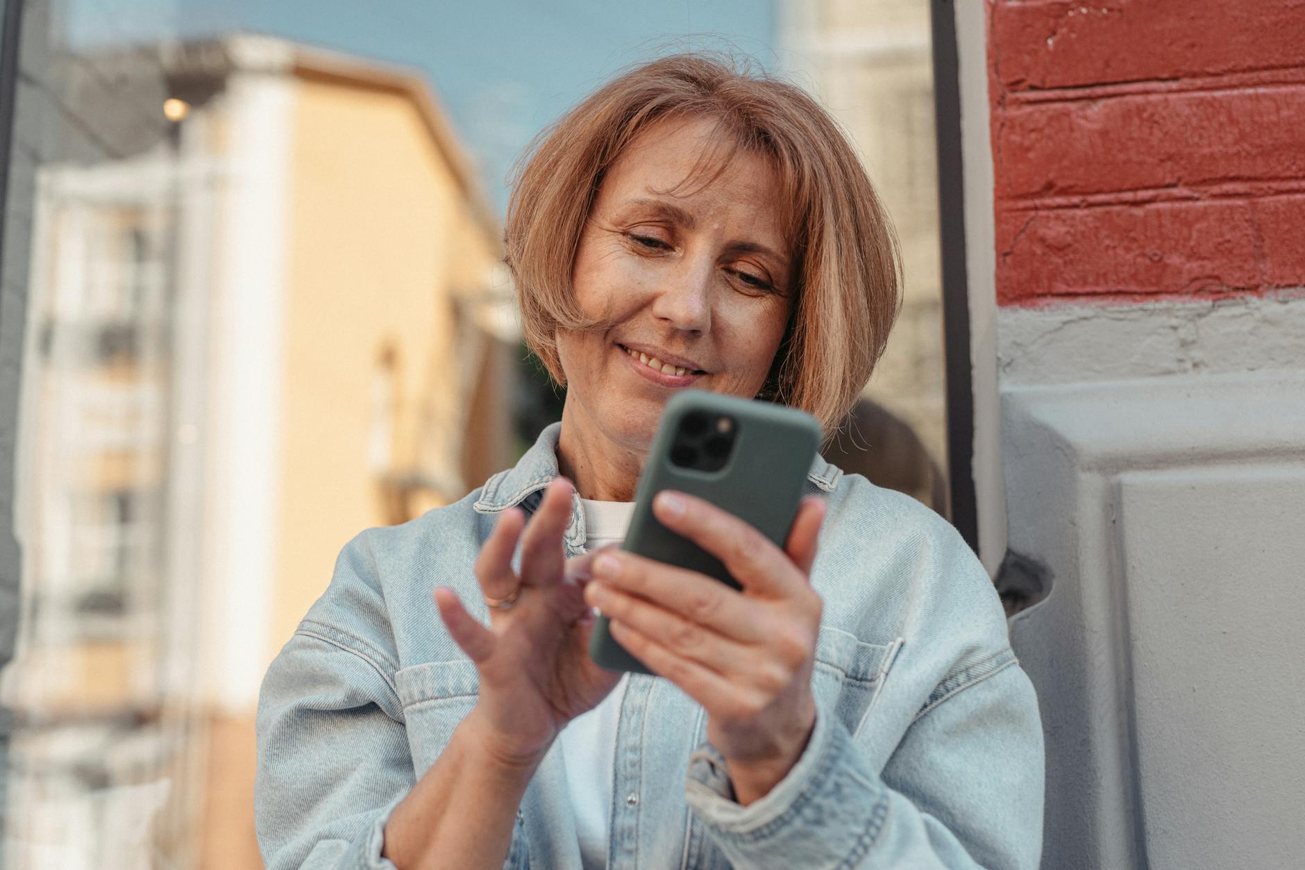 A middle-aged woman in a denim jacket smiles while using her smartphone outside.