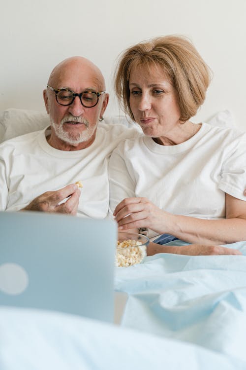 An Elderly Couple Eating Popcorn while Watching in Bed