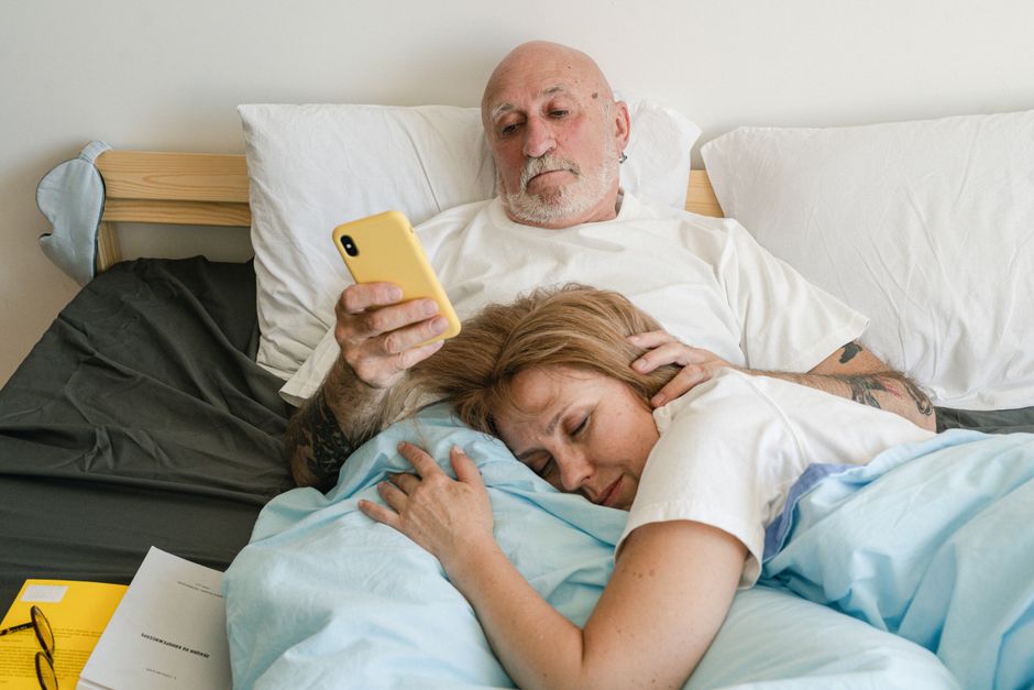 A senior man and woman relaxing in bed, with man using a smartphone while woman sleeps.