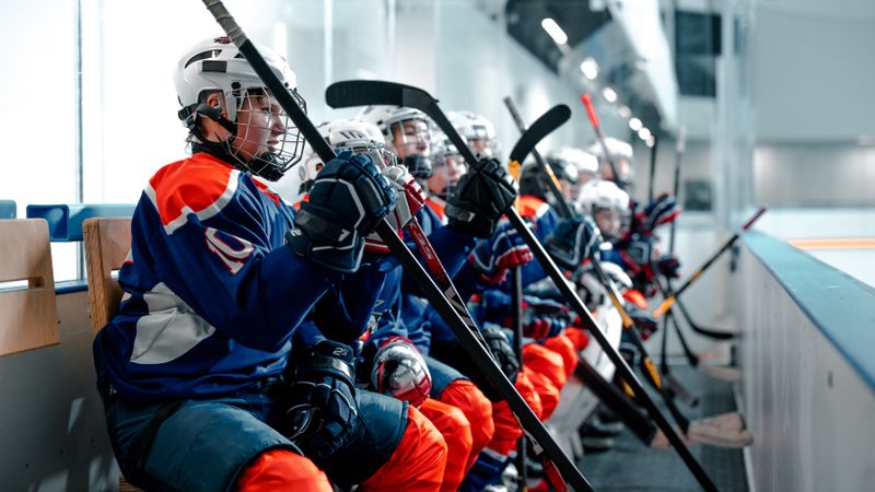 Young ice hockey players in uniforms sitting on the bench during a game, holding hockey sticks.