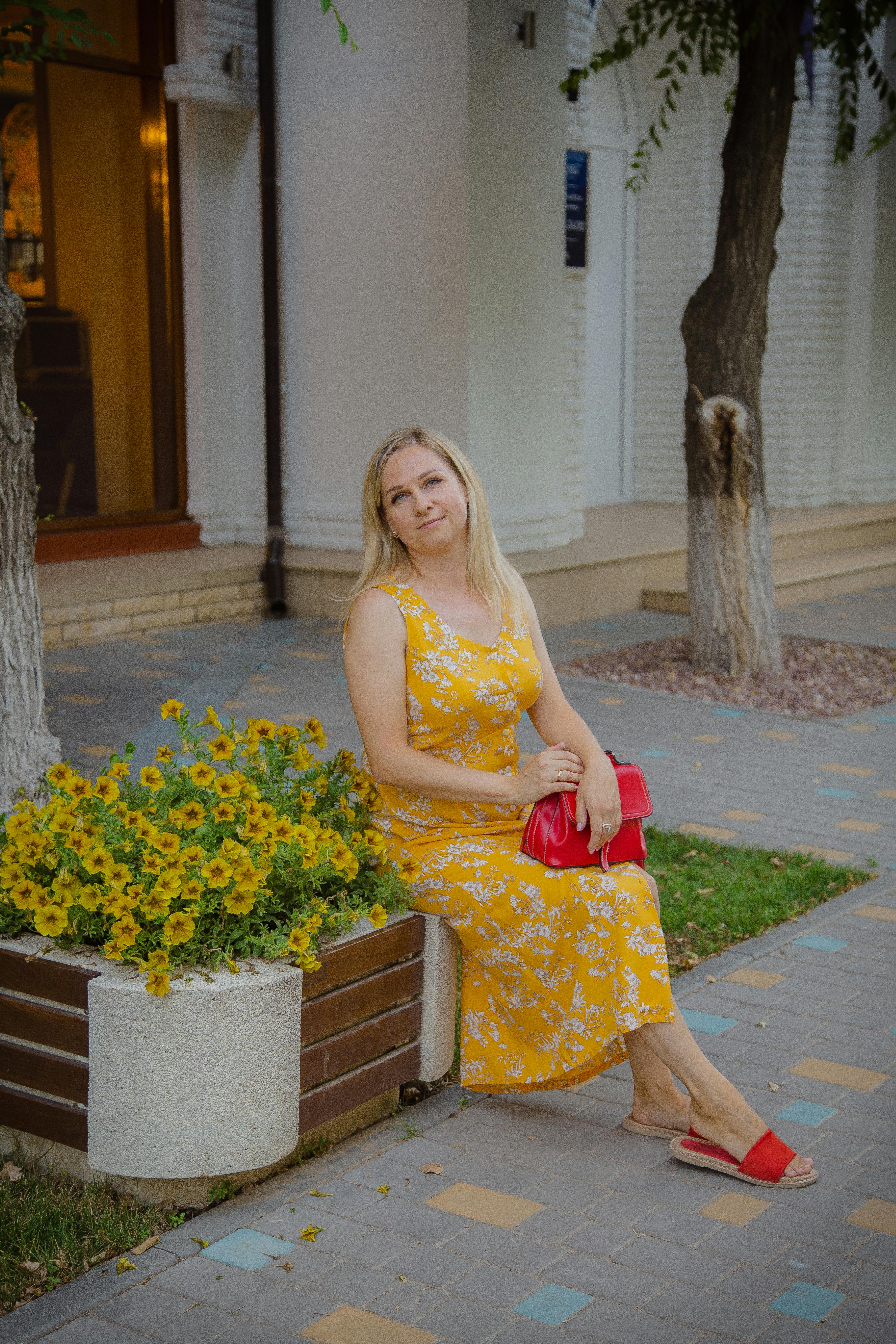Free Smiling woman in a yellow dress sits by flowers on a summer city sidewalk. Stock Photo