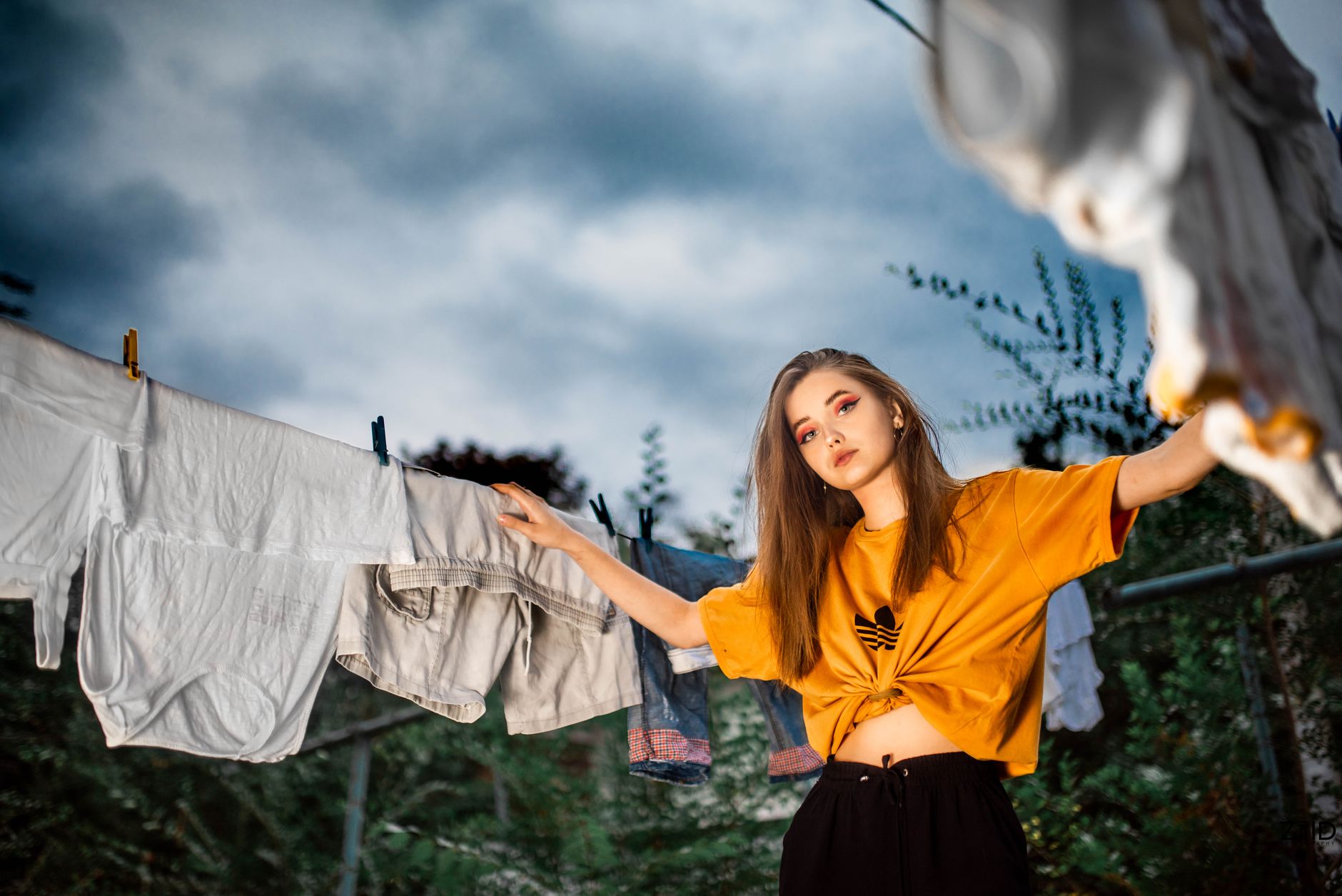 A young woman in a yellow shirt hanging clothes on a line amidst greenery and an overcast sky.