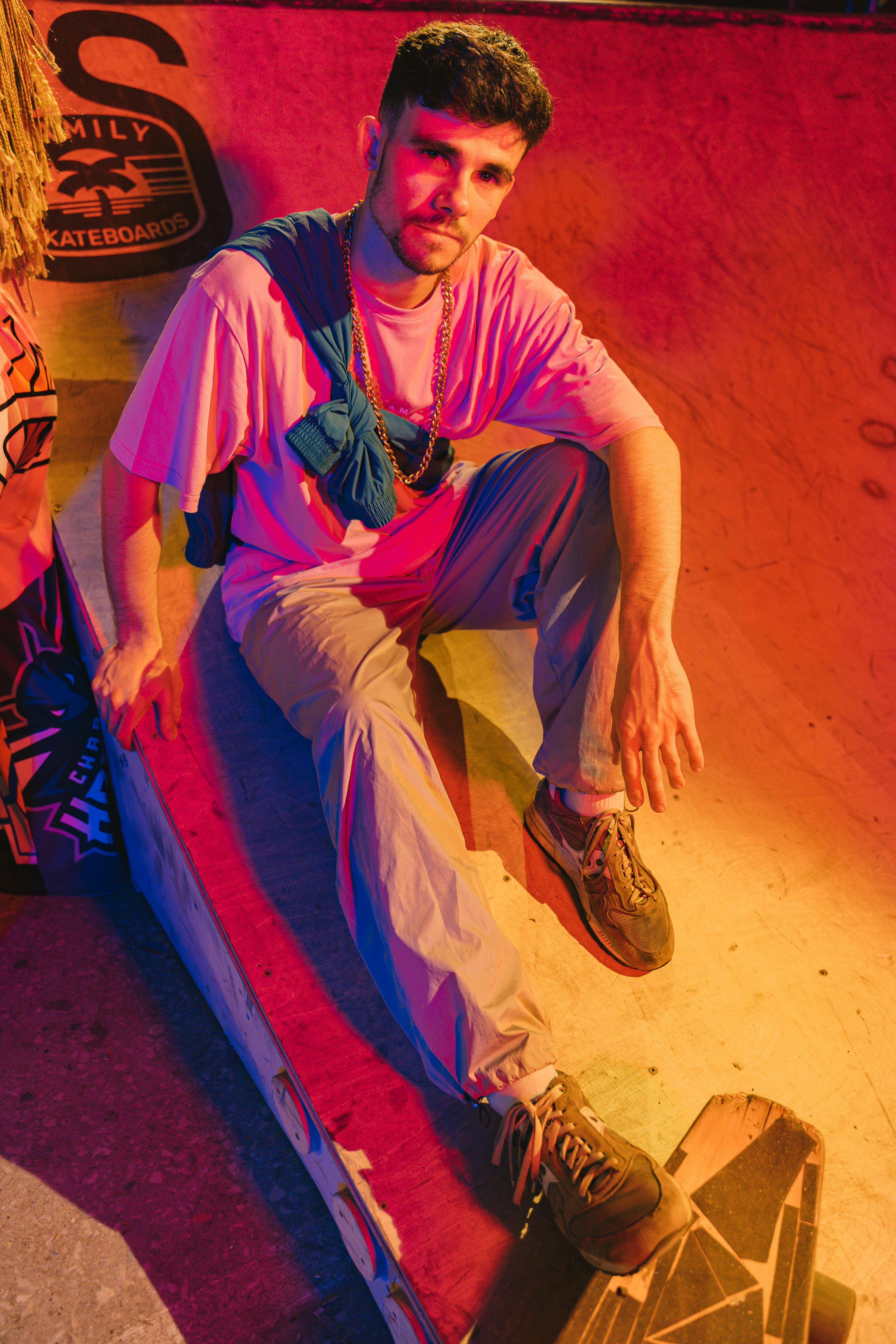A young man sits casually at an indoor skatepark, illuminated by vibrant red and yellow lights.