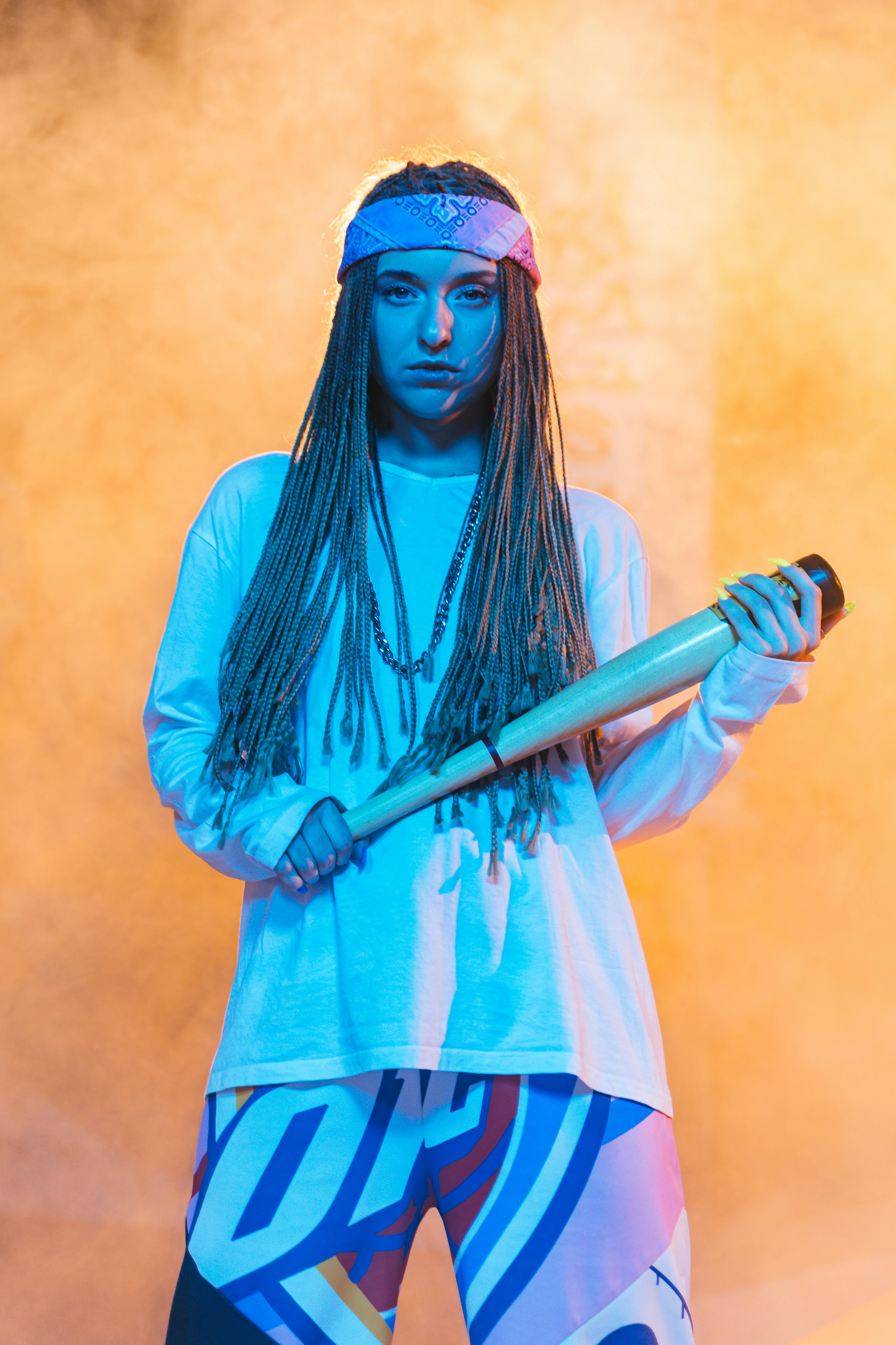 A fashionable woman with braids and a bat poses confidently against a vibrant backdrop.