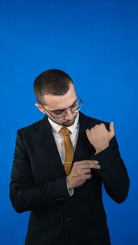 Portrait of a young man in a black suit adjusting his sleeve, set against a bright blue background.
