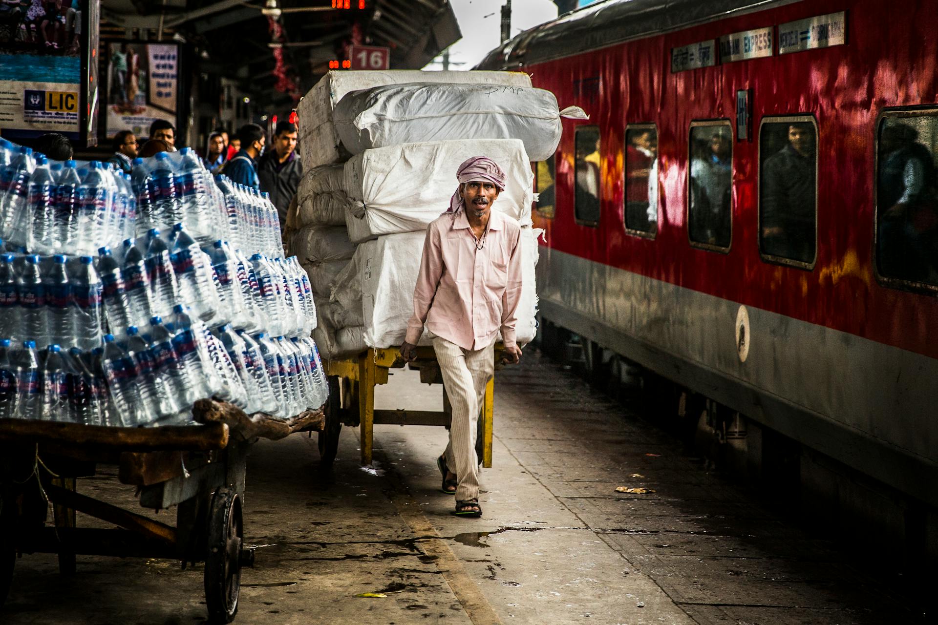 Vendor transporting heavy load beside a train with determination at a busy station.