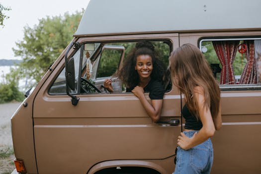 Two women smiling and bonding by a camper van on a summer road trip.