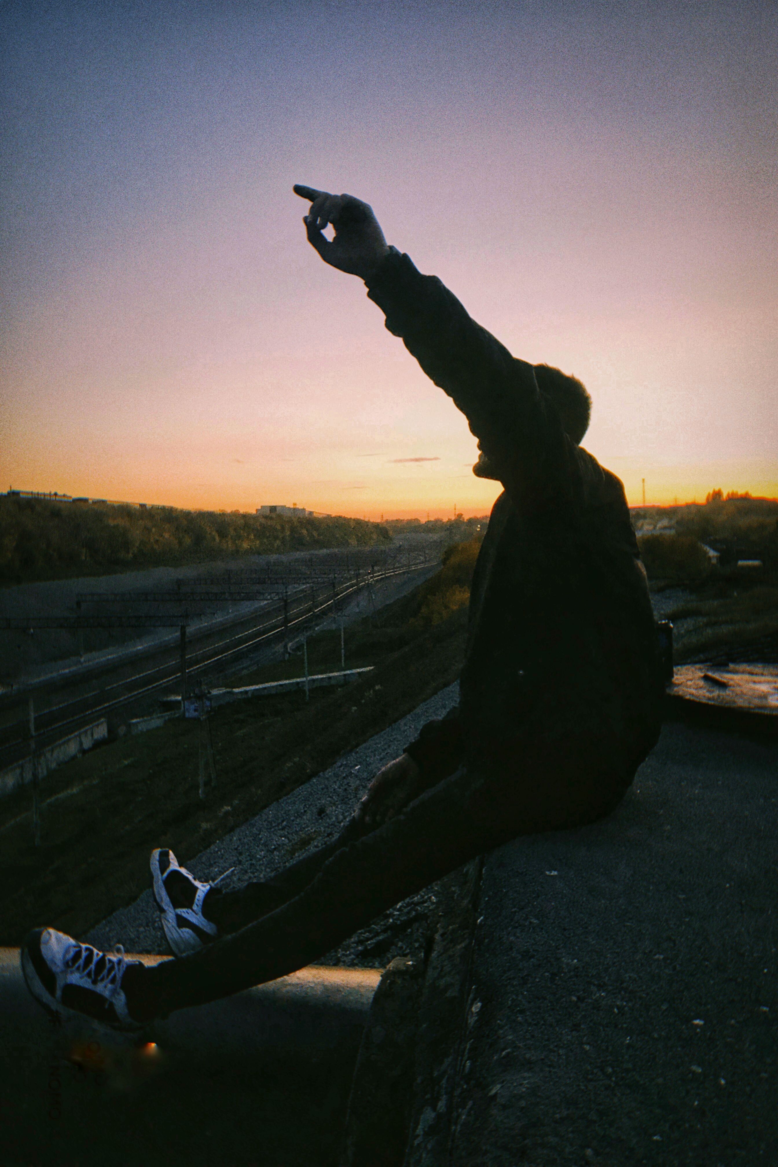 A person silhouetted against a sunset, sitting by train tracks, pointing towards the horizon.