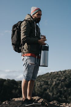 Bearded man with backpack holding a vacuum flask of mate on a mountain, embracing nature.