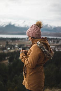 Woman wearing beanie and jacket using a smartphone outdoors with snow-capped mountains in the background.