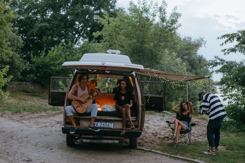 Free Group of friends relax and play guitar by a van in a serene outdoor setting, embodying the joy of summer evenings. Stock Photo