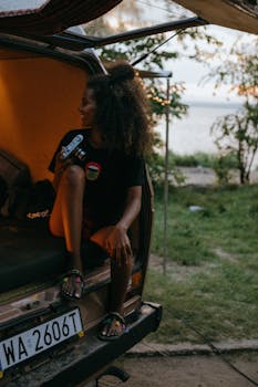 A woman with curly hair sits in a camper van holding a camera, enjoying a peaceful evening outdoors.