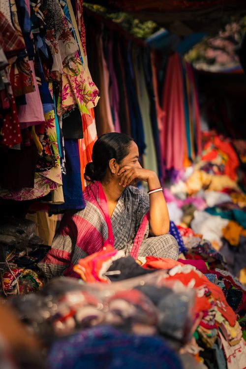Free A woman sits at a vibrant street market surrounded by colorful fabrics and clothes on display. Stock Photo