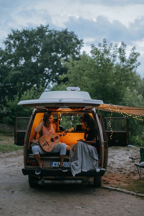 Free Two friends enjoy a cozy evening camping with a guitar by a van in the forest. Stock Photo