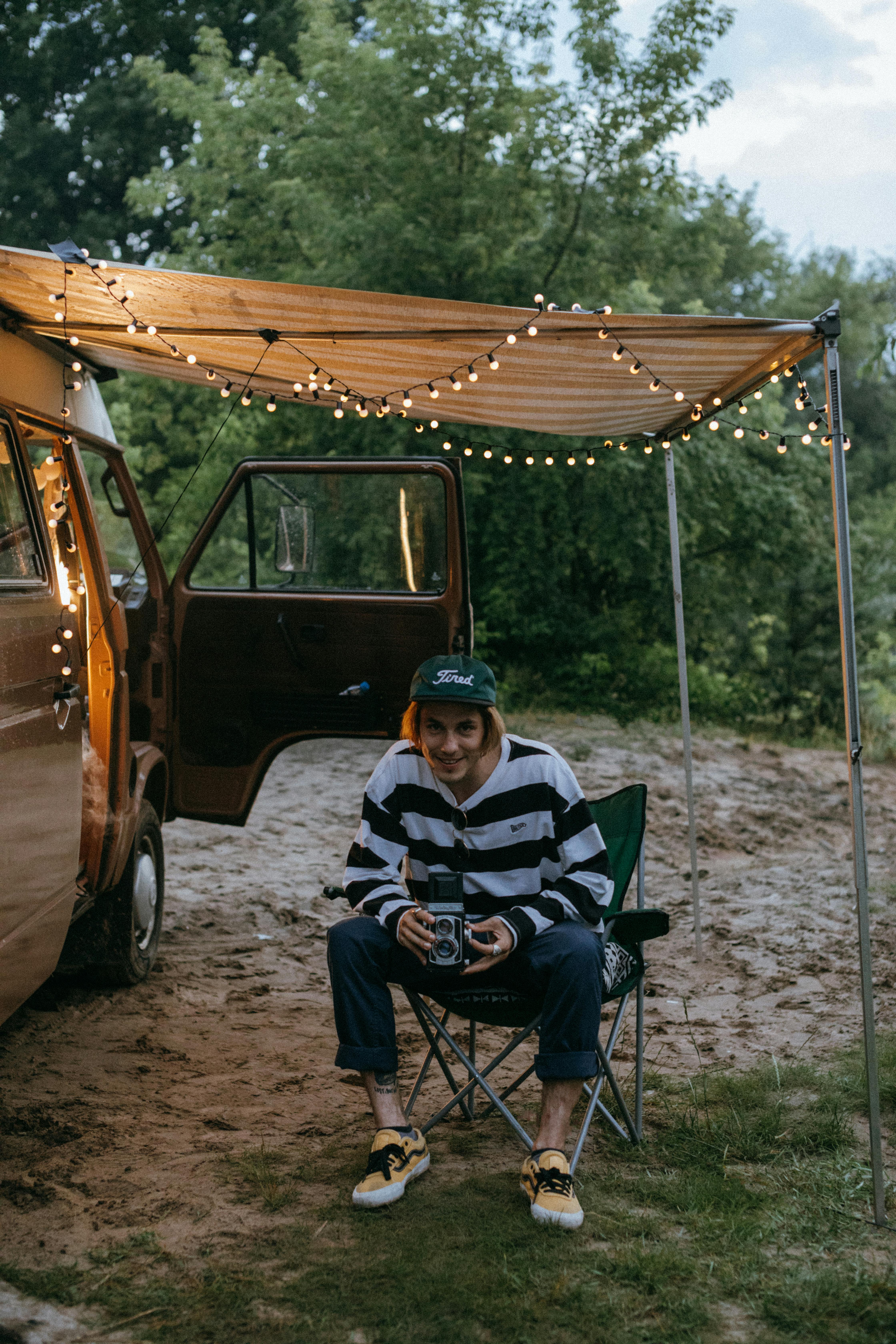 A relaxed camper enjoys dusk outdoors under twinkling lights, featuring a van and portable chair.